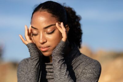 Close-up of a woman in a gray sweater holding her temples, experiencing a headache outdoors in the sunlight.
