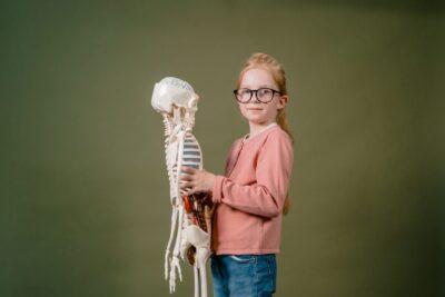 A girl in glasses holding a human skeleton model against a plain background, indoors.