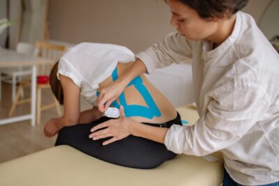 A therapist applies blue kinesiology tape to a patient's back in a clinical setting.